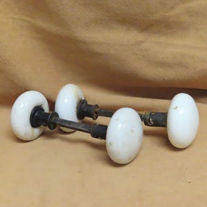 May include: Four antique white porcelain door knobs with black metal stems. The knobs have an oval shape and are arranged in a horizontal line. The background is a solid tan color.