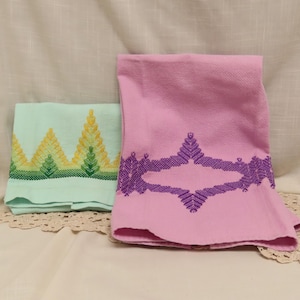 May include: A folded light green towel featuring a yellow and green geometric cross-stitch border pattern, placed next to a pink towel with a purple geometric cross-stitch design centered on the fabric, both made of textured cotton material.
