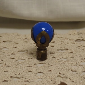 Puede incluir: Un remate de vidrio azul con detalles dorados. El remate tiene una parte superior redonda y brillante de color azul y una base decorativa dorada. La base está unida a una varilla metálica de color latón.