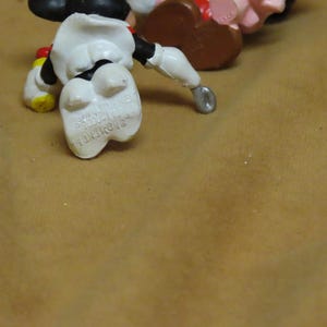 Puede incluir: Primer plano de una figura de Mickey Mouse tumbada boca arriba. La figura es principalmente blanca, con pantalones cortos negros, zapatos amarillos y guantes rojos. Las palabras "MADE IN HONG KONG" son visibles en el pie de la figura.
