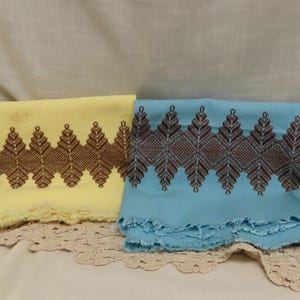 May include: Two rectangular fabric cloths, one pale yellow and one light blue, each featuring a wide horizontal band of dark brown geometric embroidery in a repeating leaf-like pattern. Both cloths have frayed, unfinished edges at the bottom.