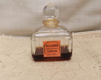 Può includere: Un flacone di profumo vintage con un tappo trasparente sfaccettato e un corpo rettangolare. Il flacone contiene un liquido ambrato scuro e presenta un'etichetta con la scritta "BELLODGIA CARON PARIS FRANCE."
