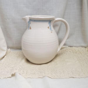 May include: A white ceramic pitcher with a rounded body and a handle. The pitcher has a blue band around the rim with small floral accents. The pitcher is sitting on a cream-colored lace doily.