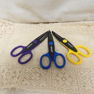 May include: Three pairs of Singer brand scissors in purple, blue, and yellow. Each pair has black blades and colored handles. The scissors are arranged on a cream-colored, lace-patterned surface.