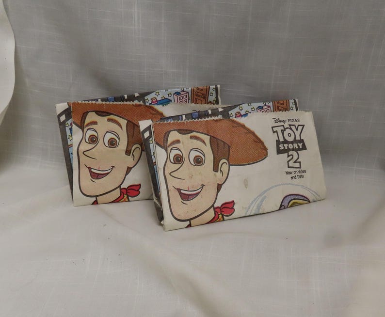 May include: Two white cardboard lunch bags with a Toy Story 2 theme. The bags feature a cartoon image of Woody from the movie. The text on the bag reads "Disney Pixar Toy Story 2 Now on Video and DVD".