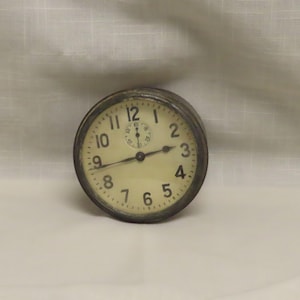 Könnte beinhalten: Eine runde Vintage-Uhr mit cremefarbenem Zifferblatt und schwarzen Ziffern. Die Uhr hat ein dunkles Metallgehäuse und ein kleineres Zifferblatt oben in der Mitte. Die Zeiger sind schwarz, und die Uhr steht auf einer weißen Oberfläche.