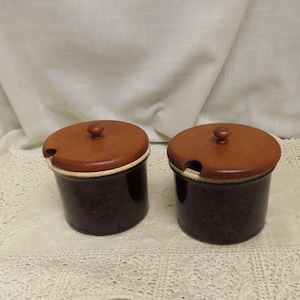 以下が含まれることがあります： 木製の蓋が付いた2つの茶色のセラミックジャー。蓋には小さなノブとスプーン用の切り欠きがあります。ジャーは白いレースのテーブルクロスの上にあります。背景は白いカーテンです。