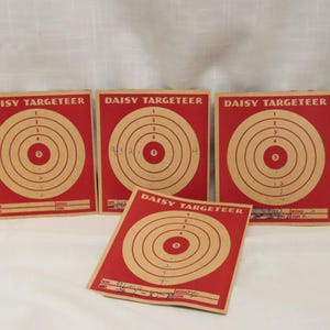 May include: Four vintage Daisy Targeteer shooting targets. Each target has a red background with concentric tan rings and numbers. The text "DAISY TARGETEER" is printed at the top of each target.