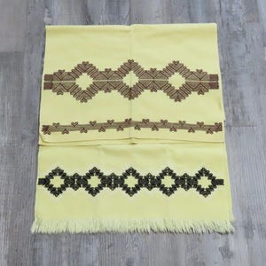 Puede incluir: Una toalla amarilla doblada con patrones geométricos bordados en marrón y negro. La toalla tiene un borde con flecos y un diseño decorativo de diamantes y corazones.