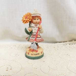 Puede incluir: Una figurita de una niña con un gorro blanco de punto y un suéter marrón que sostiene un rastrillo y un girasol. La base de la figurita es verde y dice "OCTOBER" en letras rojas.