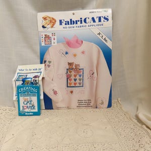 May include: A FabriCATS no-sew fabric applique kit featuring a light pink sweatshirt with embroidered cat designs, hearts, and butterflies. A small blue box with the text "CAT LOVER" is also visible.