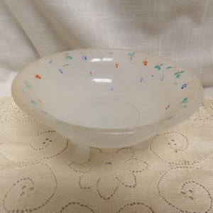 Op de afbeelding: Een witte, ovale glazen kom met drie kleine voetjes. De kom is versierd met kleine, kleurrijke bloemmotieven in rood, blauw en groen. De kom staat op een crèmekleurig, kantachtig oppervlak.