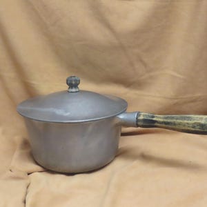May include: A small, vintage metal saucepan with a lid and wooden handle. The pot is silver-colored with a rounded body and a lid with a small knob. The handle is made of wood with a dark, weathered appearance. A small metal loop is at the end of the handle.