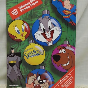 Puede incluir: Un anuncio navideño de Warner Bros. Studio Store de 1999. La imagen presenta adornos de personajes de dibujos animados, incluyendo a Piolín, Bugs Bunny, Scooby-Doo y Batman. También hay una figura de Superman.