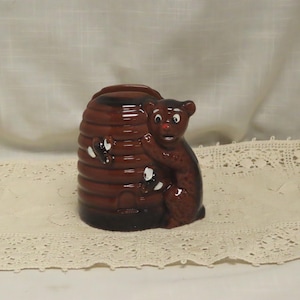 May include: A brown ceramic honey pot shaped like a beehive with a bear clinging to the side. The beehive has white accents and the bear has a surprised expression. The pot is sitting on a lace doily.