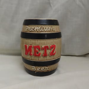 Op de afbeelding: Een keramische bierkan in de vorm van een vat met zwarte banden en de woorden "Premium Metz Beer" in rode en witte letters.