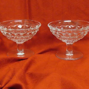 May include: Two clear, stemmed champagne coupe glasses with diamond-cut patterned bowls. The glasses sit on a red-orange fabric background. The glasses are made of glass.