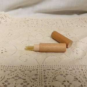 May include: A wooden needle case with a removable lid, partially open to reveal a bundle of needles. The case and lid are made of light-colored wood. The needles are thin and gold-colored. The case is resting on a cream-colored lace tablecloth.