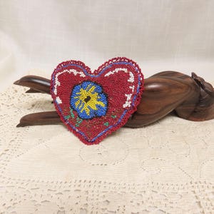 May include: A red beaded heart with a blue and yellow flower design. The heart is lying on a wooden surface.