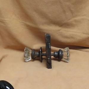 May include: Antique door hardware set featuring a dark metal latch and two clear glass doorknobs. The glass knobs have a ribbed design. The set is designed for a vintage or rustic aesthetic.