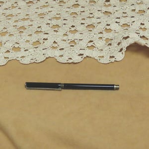 May include: A black pen with a silver clip and tip lies on a tan surface. A portion of a cream-colored, crocheted lace fabric is visible in the background.
