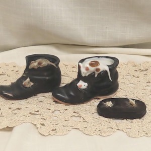May include: Two black ceramic shoe-shaped containers and a separate black oval lid. The shoes feature small sculpted mice in white, brown, and tan colors perched on the sides and rims. The items have a smooth finish and visible mouse details.