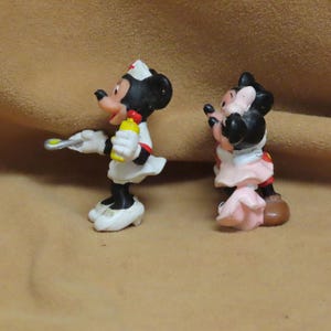 Puede incluir: Dos figuras de Minnie Mouse. Una est&aacute; vestida de enfermera con uniforme blanco y cruz roja en la gorra, sosteniendo una botella amarilla y una jeringa. La otra lleva un vestido rosa y zapatos rojos.