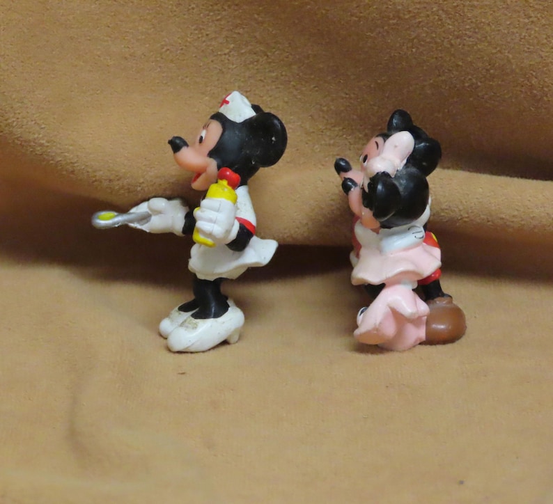 Puede incluir: Dos figuras de Minnie Mouse. Una est&aacute; vestida de enfermera con uniforme blanco y cruz roja en la gorra, sosteniendo una botella amarilla y una jeringa. La otra lleva un vestido rosa y zapatos rojos.
