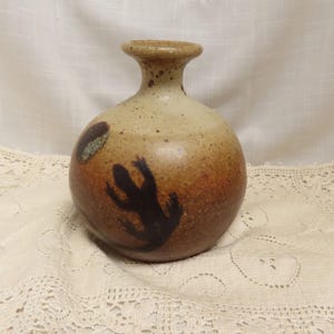 May include: A handcrafted ceramic vase with a rounded body and a narrow neck. The vase features a speckled tan and brown glaze with a dark brown lizard silhouette design. The vase is a decorative item.