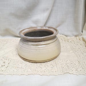 May include: A handmade ceramic pot with a wide opening and a rounded body. The pot features a light beige exterior with subtle brown and gray accents around the rim. The interior is dark. The pot is likely used for decorative purposes.