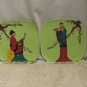Puede incluir: Dos platos cuadrados de cerámica con fondo verde claro. Cada plato presenta una figura asiática pintada bajo una rama de árbol. Uno toca un instrumento de cuerda, el otro una flauta. Las figuras visten coloridas túnicas.
