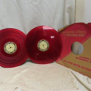 May include: Two vintage red Recordo Discs with a gold and black label, next to a red and tan record sleeve. The sleeve reads "Recordo Discs by Wilcox-Gay" and "Aluminum Base Professional Quality".
