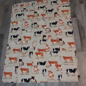 Op de afbeelding: Een beige quilt met een herhaald patroon van bruine, zwarte en witte koeien. De quilt heeft een gefranjerde rand, wat suggereert dat hij handgemaakt is. De koeien staan in verschillende poses, wat een rustiek, boerderij-thema ontwerp creëert.