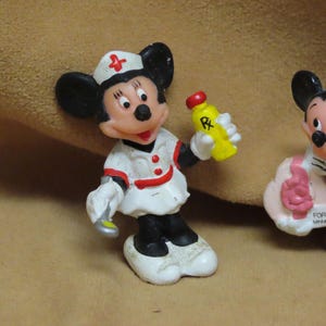 Puede incluir: Una figura vintage de Mickey Mouse vestida de enfermera, sosteniendo un frasco de medicina amarillo con una tapa roja. La figura lleva un uniforme blanco con detalles rojos y un gorro de enfermera con una cruz roja. Otra figura de Mickey Mouse es visible en el fondo.