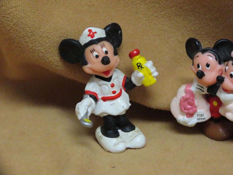 Puede incluir: Una figura vintage de Mickey Mouse vestida de enfermera, sosteniendo un frasco de medicina amarillo con una tapa roja. La figura lleva un uniforme blanco con detalles rojos y un gorro de enfermera con una cruz roja. Otra figura de Mickey Mouse es visible en el fondo.