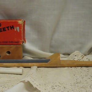 May include: A vintage wooden knife sharpener with a rectangular sharpening stone. The sharpener is accompanied by a red box with the text "Colorado Ever Teeth". The background is a white cloth.