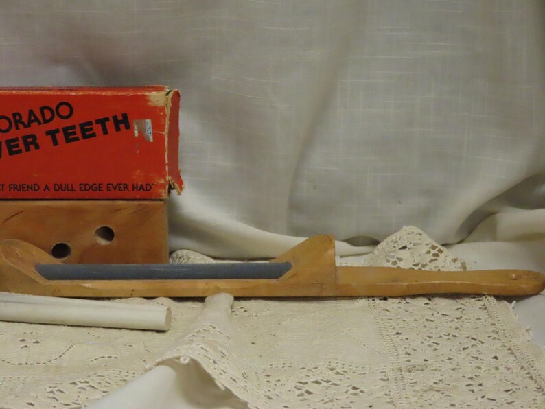 May include: A vintage wooden knife sharpener with a rectangular sharpening stone. The sharpener is accompanied by a red box with the text "Colorado Ever Teeth". The background is a white cloth.