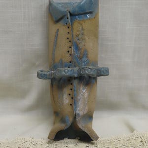May include: A ceramic vase shaped like a shirt, featuring a light blue collar and accents. The vase has a tan body with blue floral designs and a decorative band. The design includes button details and a unique, artistic aesthetic.