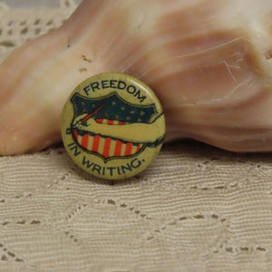 May include: A vintage button with the words "Freedom in Writing" in a circular design. The button features a shield with stars and stripes, and a hand holding a pen. The button is set against a seashell and lace.