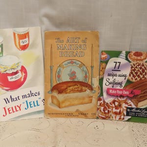 May include: Three vintage cookbooks on a white surface. The books feature titles like "What makes JELLY JELL?" and "The Art of Making Bread." The third book is titled "77 recipes using Swiftning Make Your Own Mix."