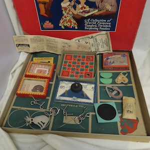 May include: Vintage puzzle game set with a red lid featuring the text "A Collection of World Famous Tangled, Twisted, Perplexing Puzzles." The set includes various puzzles, including a square problem, a board game, and metal wire puzzles.