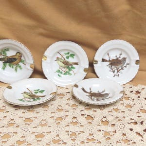 May include: Five white ceramic ashtrays with gold trim and bird illustrations. Each ashtray features a different bird design in shades of brown, green, and yellow. The ashtrays are arranged on a beige surface.
