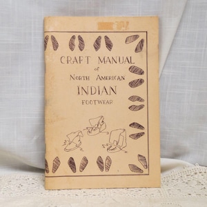 May include: A vintage craft manual titled "Craft Manual of North American Indian Footwear." The cover is tan with brown illustrations of footprints and footwear. The manual appears aged, with a worn appearance.