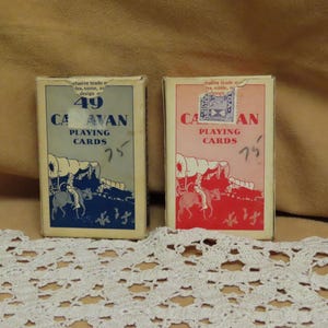 May include: Two vintage Caravan playing card decks. One deck has a blue and gray design, the other a pink and red design. Both feature a covered wagon illustration and the text "Caravan Playing Cards". The price "75" is written on each deck.