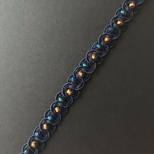 May include: A dark blue beaded trim with a repeating pattern. The trim features alternating iridescent teal and bronze-colored beads, woven into a dark blue cord. The design is suitable for embellishing clothing or accessories.