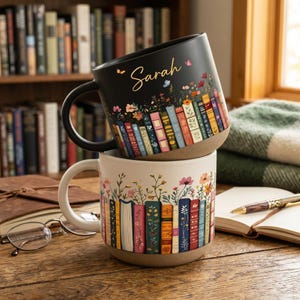 Op de afbeelding: Twee keramische mokken, één zwart en één crème, gestapeld op een houten tafel. De zwarte mok heeft de naam "Sarah" en een ontwerp van boeken en bloemen. De crèmekleurige mok heeft een vergelijkbaar ontwerp. Op de achtergrond een boekenkast.