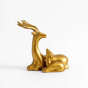 May include: A gold-coloured deer ornament featuring a mother and fawn. The mother deer stands tall, while the fawn rests. The ornament has a smooth, reflective finish and is set against a white backdrop. Suitable for home decoration, this piece adds a touch of elegance.