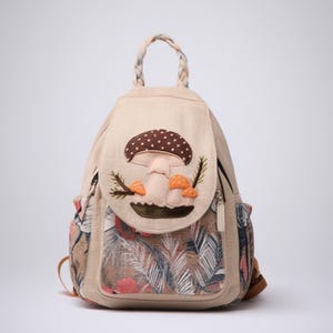 Op de afbeelding: Beige rugzak met een paddenstoel applicatie. De flap heeft een grote paddenstoel met bruine stippen en kleinere oranje paddenstoelen. De zijkanten hebben bloemen- en bladerpatronen. De tas heeft een gevlochten handvat en twee zijzakken.