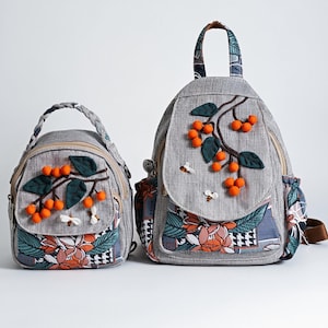 May include: Two grey backpacks with floral and fruit embroidery. The larger backpack has a top handle and adjustable straps. The smaller backpack has a braided handle. Both feature orange pom-poms, green leaves, and bee appliqués.