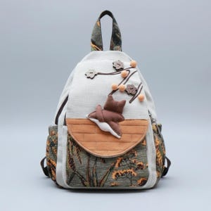 Może przedstawiać: Kremowy plecak z kwiatowym wzorem. Plecak posiada brązową aplikację w kształcie gwiazdy oraz kieszeń z klapką z brązowo-pomarańczowym wzorem. Uchwyt i boczne kieszenie mają kwiatowy wzór.